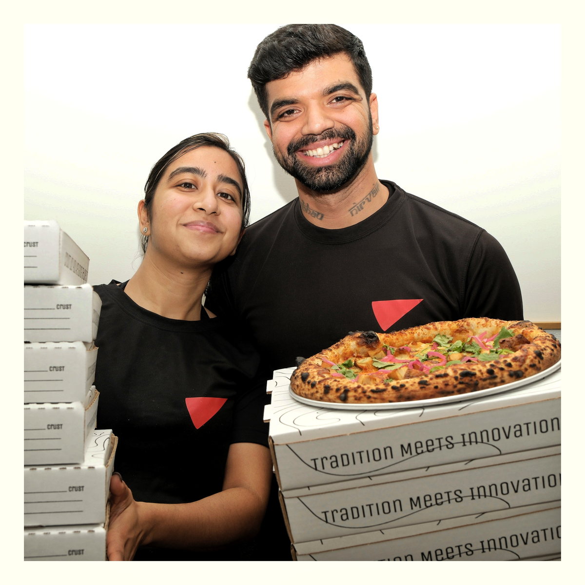 Chef Sohini and Mayur, the couple behind Baked Columbia Pizza, holding a stack of pizza boxes and a freshly baked pie